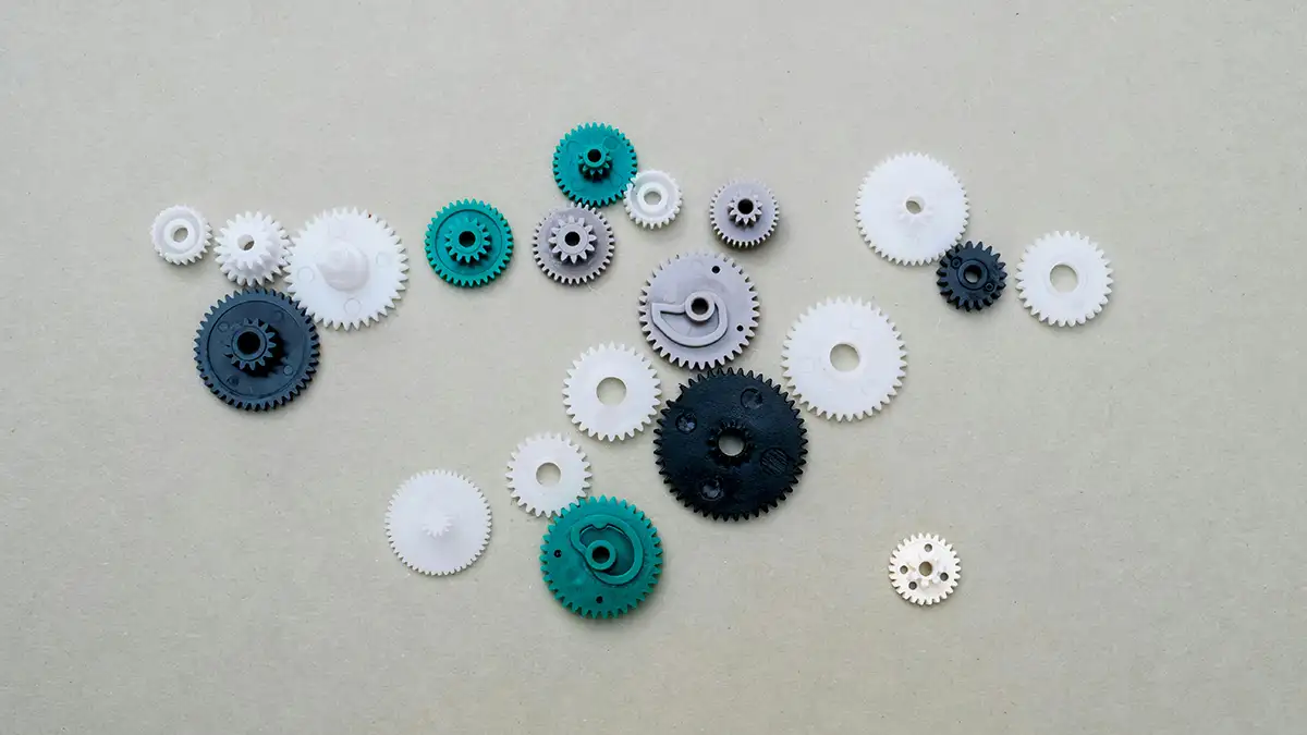 Assorted plastic gears in various sizes and colors, including white, black, green, and gray, commonly used in precision mechanisms made of POM and other engineering plastics
