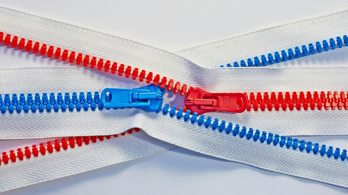 Close-up of red and blue plastic zippers with white fabric tape, showing interlocking teeth and POM zipper sliders in a crisscross pattern.